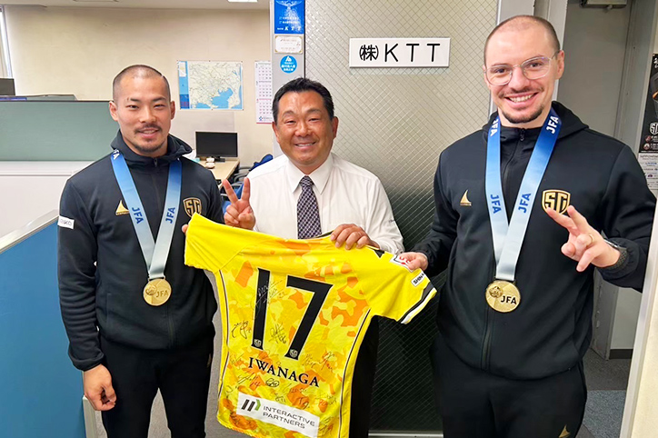 SHINAGAWA CITY FUTSAL CLUBの選手が会社訪問。選手に挟まれてユニフォームをもってVサインをする橋本代表