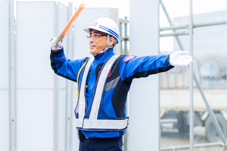 道路に面した工事現場入り口で、ヘルメットを被った警備員が工事用車両を誘導している様子